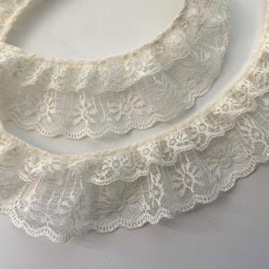 May include: Two pieces of ivory lace trim with a scalloped edge. The lace has a floral pattern and is layered, creating a ruffled effect. The trim is likely used for sewing or crafting projects.