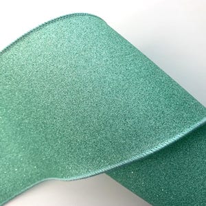 May include: A close-up of a roll of teal-colored ribbon with a glittery texture. The ribbon has a stitched edge and is partially rolled, revealing its texture and color. The background is white.