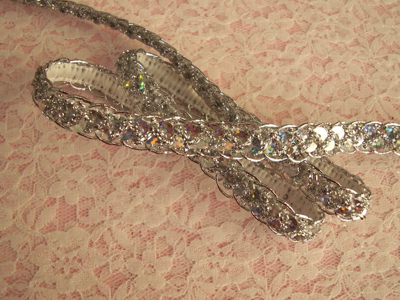 May include: Silver sequined trim with iridescent sequins. The trim is woven and has a raised texture.
