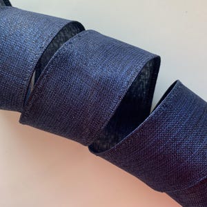 May include: A roll of navy blue ribbon with a textured weave. The ribbon is spiraled, showcasing its width and the subtle sheen of the fabric. The close-up shot highlights the detailed texture and color of the decorative ribbon.