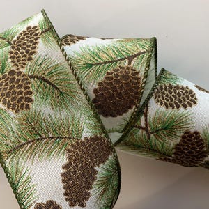 May include: A decorative ribbon featuring a repeating pattern of brown pine cones and green pine needles on a white background. The ribbon has a green border and gold glitter accents, perfect for holiday crafts and decorations.