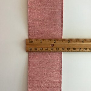 May include: A close-up of a pink ribbon with a shimmery finish. A ruler is placed next to the ribbon to show its width, which is 1 inch.