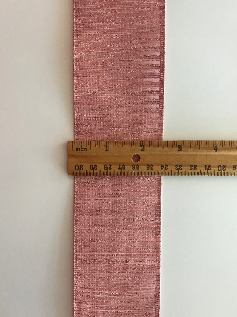 May include: A close-up of a pink ribbon with a shimmery finish. A ruler is placed next to the ribbon to show its width, which is 1 inch.