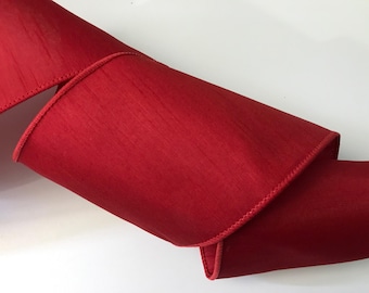 May include: A length of rich red ribbon, displaying a slight sheen. The ribbon is folded and curved, highlighting its texture and the stitched edge. The material appears to be a smooth fabric, suitable for crafting or gift wrapping.