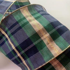 May include: A wide fabric ribbon featuring a plaid pattern in shades of navy blue, forest green, and cream. The ribbon has a metallic gold thread woven into the plaid design and is finished with a thin, flexible gold wire along both edges.