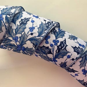 May include: A rolled-up ribbon with a repeating pattern of blue holly leaves and berries on a white background. The ribbon is made of a textured fabric and is suitable for crafts and holiday decorations.