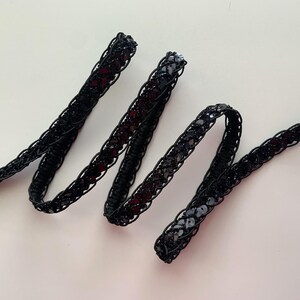 May include: Black sequined trim with a braided design. The trim is made of a black fabric with small, round sequins.