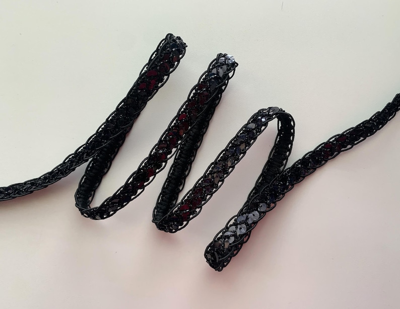 May include: Black sequined trim with a braided design. The trim is made of a black fabric with small, round sequins.