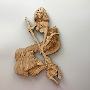 May include: A wooden carving of a mermaid holding a trident with an octopus on the end. The mermaid has long flowing hair and is wearing a seashell bra. The carving is painted in shades of brown.