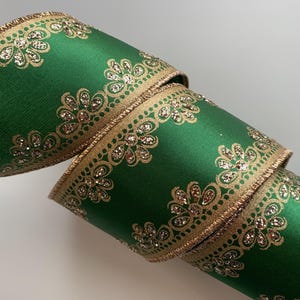 May include: A roll of emerald green ribbon with a gold floral pattern and gold trim. The ribbon is made of a satin-like material and is rolled up, showing the intricate design. The ribbon is approximately 2 inches wide.