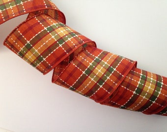 May include: A rolled-up, wide ribbon with a plaid pattern in shades of orange, red, yellow, and green. The plaid design features white stitching. The ribbon is likely used for crafting, gift wrapping, or seasonal decorations.
