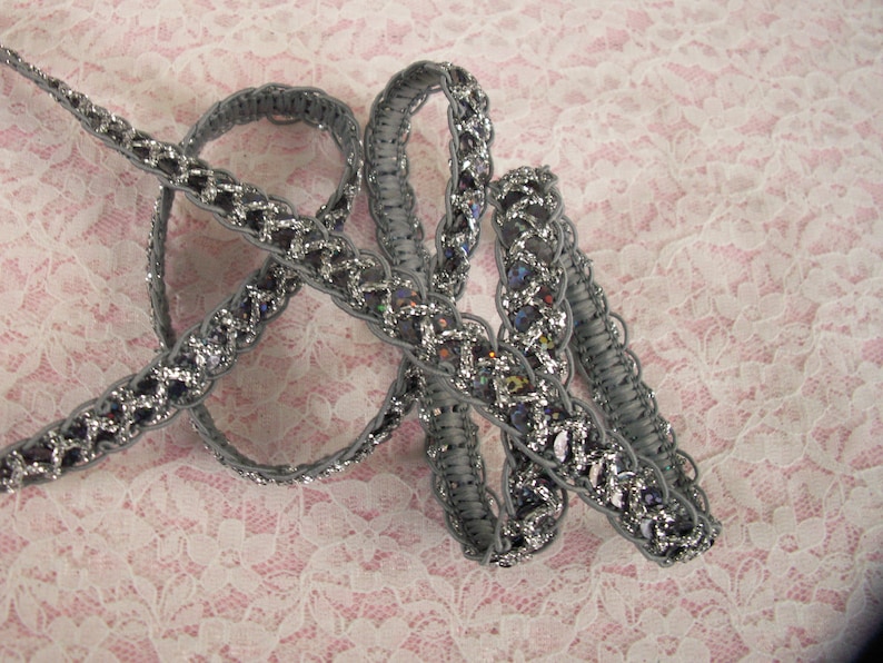 May include: Silver and gray braided trim with a sparkly finish. The trim is made of a woven fabric with a delicate pattern.