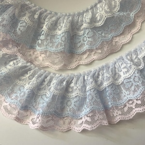 May include: Close-up of layered lace trim in pastel colors. The trim features a scalloped edge and is in shades of white, light blue, and pale pink. The delicate lace has a floral pattern, suitable for sewing and craft projects.