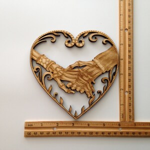 May include: Wooden heart-shaped wall decor with two skeletal hands holding each other. The hands are intricately carved and detailed.
