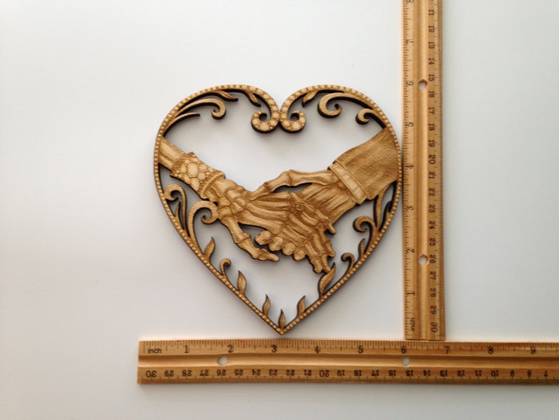 May include: Wooden heart-shaped wall decor with two skeletal hands holding each other. The hands are intricately carved and detailed.