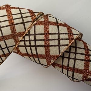May include: A decorative ribbon with a beige background and a brown plaid pattern. The plaid lines are outlined with a glittery copper color. The ribbon is rolled up, showing the pattern and texture.
