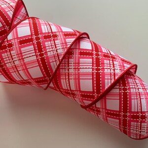 May include: A roll of decorative ribbon with a plaid pattern in shades of red, pink, and white. The ribbon has a stitched edge and is tightly coiled, showcasing the repeating design. Ideal for crafting and gift wrapping.