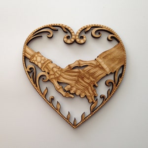 May include: A wooden heart-shaped ornament with a detailed design of two skeletal hands holding each other. The hands are intricately carved and have a natural wood finish.