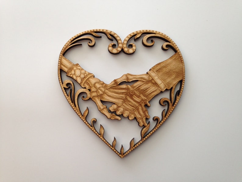 May include: A wooden heart-shaped ornament with a detailed design of two skeletal hands holding each other. The hands are intricately carved and have a natural wood finish.