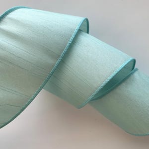 May include: A roll of light blue ribbon with a textured appearance. The ribbon has a stitched edge in a matching shade of blue. The ribbon is rolled in a spiral shape.