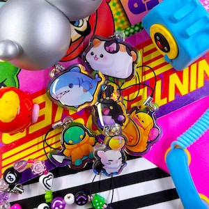 May include: A collection of colorful acrylic charms featuring various cartoon characters, including a shark, cat, and robot. The charms are attached to keychains and are set against a vibrant background with other toys and accessories.