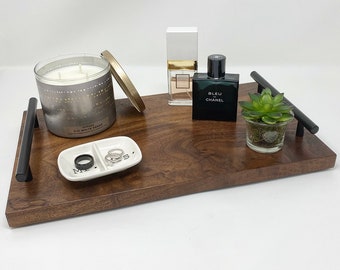 May include: A dark brown wooden tray with black handles, holding a candle, perfume bottles, a small succulent, and a ring dish with rings. The ring dish has the words "Mr" and "Mrs" on it.