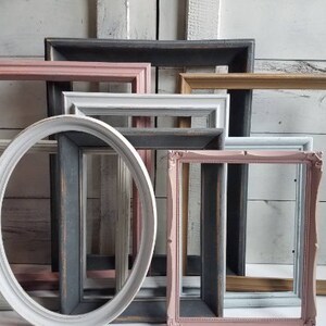 May include: A collection of empty picture frames in various shapes and colors. The frames include rectangular and oval designs, painted in shades of pink, white, gray, and gold. The frames are arranged against a white wooden background.