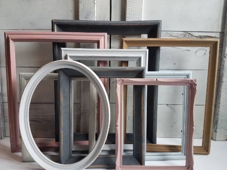 May include: A collection of empty picture frames in various shapes and colors. The frames include rectangular and oval designs, painted in shades of pink, white, gray, and gold. The frames are arranged against a white wooden background.