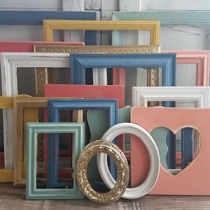 May include: A collection of various colorful picture frames in shades of blue, pink, yellow, and white. The frames are in different shapes and sizes, some with ornate details and heart cutouts. The frames are arranged against a white wooden background.