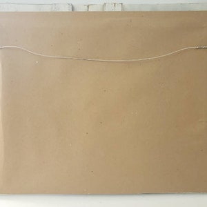 May include: Back of a framed artwork, showing a brown paper backing. A thin wire is strung across the back, secured by metal hardware for hanging. The frame is sealed with clear plastic.