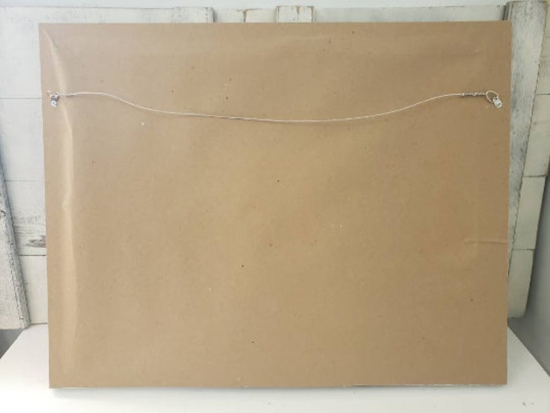 May include: Back of a framed artwork, showing a brown paper backing. A thin wire is strung across the back, secured by metal hardware for hanging. The frame is sealed with clear plastic.