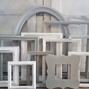 May include: A collection of various empty picture frames in shades of white and gray, arranged against a white wooden background. The frames vary in shape, including square, rectangular, and arched designs, with distressed finishes.