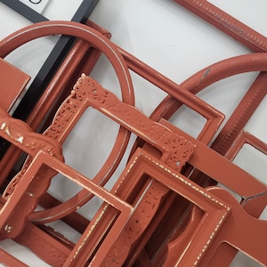 May include: A collection of various terracotta-colored picture frames, including rectangular, arched, and ornate designs. Some frames have distressed white accents, creating a vintage aesthetic. The frames are arranged on a white surface.