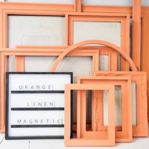 May include: A collection of various orange-colored picture frames in different shapes and sizes. A black framed sign displays the words "ORANGE LINEN MAGNETIC" in white letters. The frames are arranged against a neutral background.