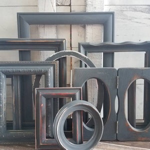May include: A collection of various dark gray picture frames in different shapes and sizes. Some frames have ornate details, while others are simple. The frames are arranged against a white wooden background, creating a rustic aesthetic.