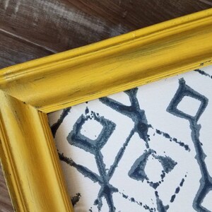 May include: A framed print with a yellow, distressed-look frame. The artwork features a white background with a repeating pattern of dark blue diamond shapes. The frame's corner is visible, showing the joinery and texture.