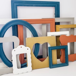 May include: A collection of various painted wooden picture frames in different shapes and sizes. The frames are in shades of blue, white, orange, gray, and yellow. Some frames have ornate details, while others are simple.