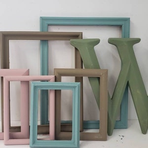 May include: A collection of decorative picture frames in various sizes and colors, including pink, teal, and brown. A large, green wooden letter "W" is also present, suggesting a home decor theme.