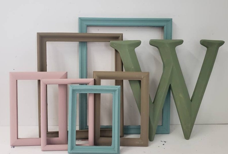 May include: A collection of decorative picture frames in various sizes and colors, including pink, teal, and brown. A large, green wooden letter "W" is also present, suggesting a home decor theme.