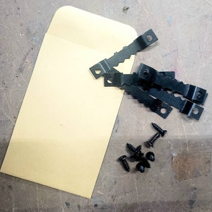 May include: A collection of black sawtooth picture hangers, screws, and a tan envelope. The hangers have a zigzag edge and a hole for mounting. The screws are small and black. The envelope is rectangular.