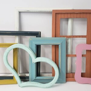 May include: A collection of colorful, empty picture frames in various shapes and sizes. The frames are painted in shades of yellow, blue, pink, white, and brown. One frame is heart-shaped. The frames have a distressed, vintage look.