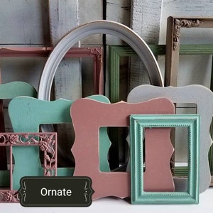 May include: A collection of colorful, empty picture frames in various shapes and sizes. The frames are painted in shades of pink, green, and white. One frame has the word "Ornate" on it. The frames are arranged in front of a light blue background.