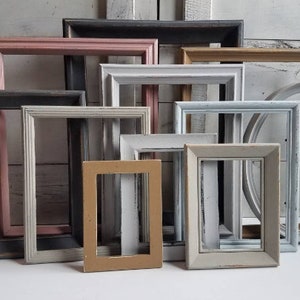 May include: A collection of various-sized picture frames in a variety of colors, including pink, gray, white, and tan. Some frames are rectangular, and one is oval. The frames have a distressed, vintage appearance.