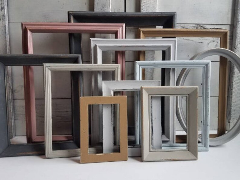May include: A collection of various-sized picture frames in a variety of colors, including pink, gray, white, and tan. Some frames are rectangular, and one is oval. The frames have a distressed, vintage appearance.