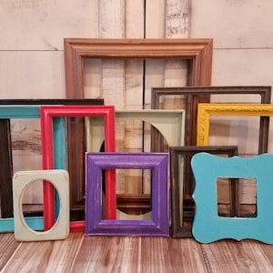 May include: A collection of colorful, empty picture frames in various shapes and sizes. The frames are painted in shades of teal, red, yellow, purple, and brown. Some frames have ornate details, while others are simple and rectangular. The frames are arranged on a wooden surface.