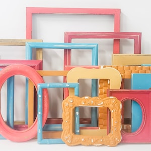 May include: A collection of colorful picture frames in various shapes and sizes. The frames are painted in shades of pink, blue, yellow, and orange. Some frames are rectangular, oval, or have heart-shaped cutouts. The frames are arranged against a white background.