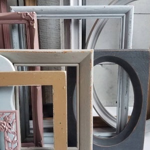 May include: A collection of various wooden picture frames in different shapes and sizes. The frames are painted in a variety of colors, including pink, blue, gray, and tan. Some frames have ornate details, while others are simple.
