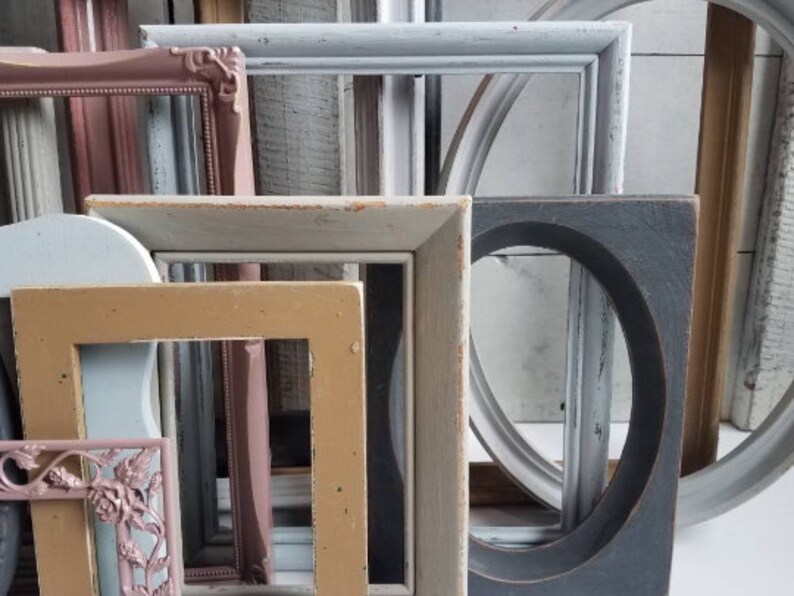 May include: A collection of various wooden picture frames in different shapes and sizes. The frames are painted in a variety of colors, including pink, blue, gray, and tan. Some frames have ornate details, while others are simple.