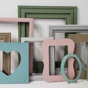 May include: A collection of painted wooden picture frames in various shapes and colors. Frames include rectangular, square, and oval designs, with colors like pink, green, blue, and gray. One frame has a heart-shaped cutout.