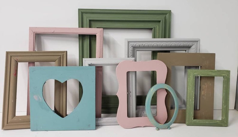 May include: A collection of painted wooden picture frames in various shapes and colors. Frames include rectangular, square, and oval designs, with colors like pink, green, blue, and gray. One frame has a heart-shaped cutout.